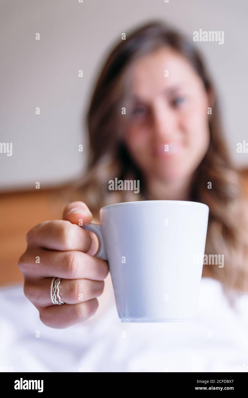 Blurred blond girl smiling to the camera holding a cup of coffee on a bed in the morning Stock Photo