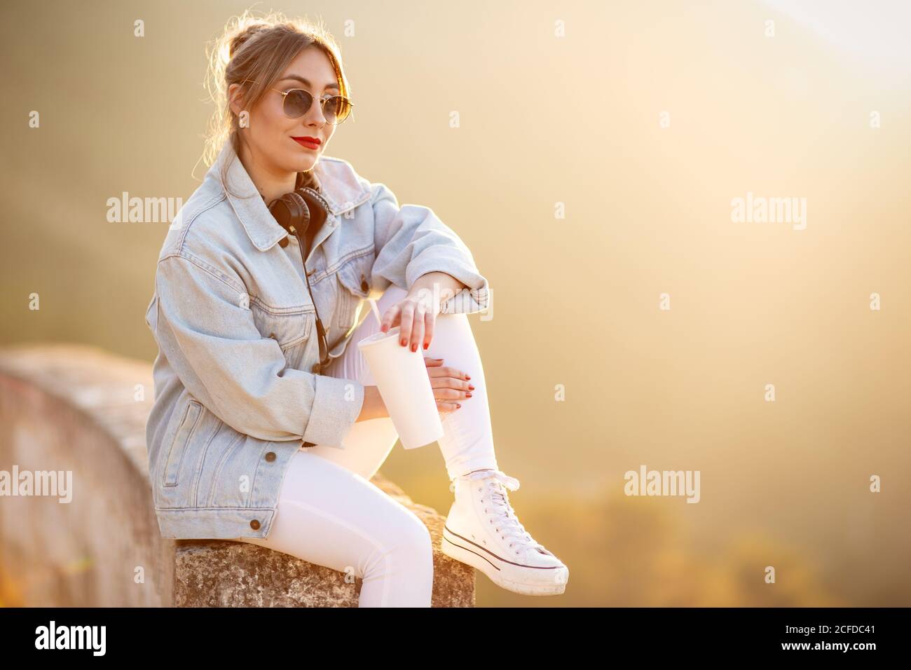 Satisfied blond haired lady in trendy sunglasses and casual wear sitting on rocky fence and looking at camera in sunlight on blurred background Stock Photo