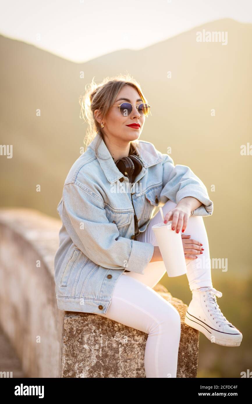 Satisfied blond haired lady in trendy sunglasses and casual wear sitting on rocky fence and looking away in sunlight on blurred background Stock Photo