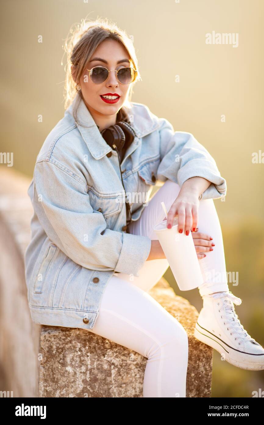 Satisfied blond haired lady in trendy sunglasses and casual wear sitting on rocky fence and looking at camera in sunlight on blurred background Stock Photo