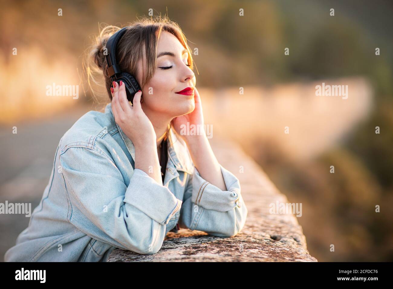 Side view of satisfied young lady in stylish casual wear and red lips leaning elbow on rocky fence with closed eyes while listening music with headphones Stock Photo