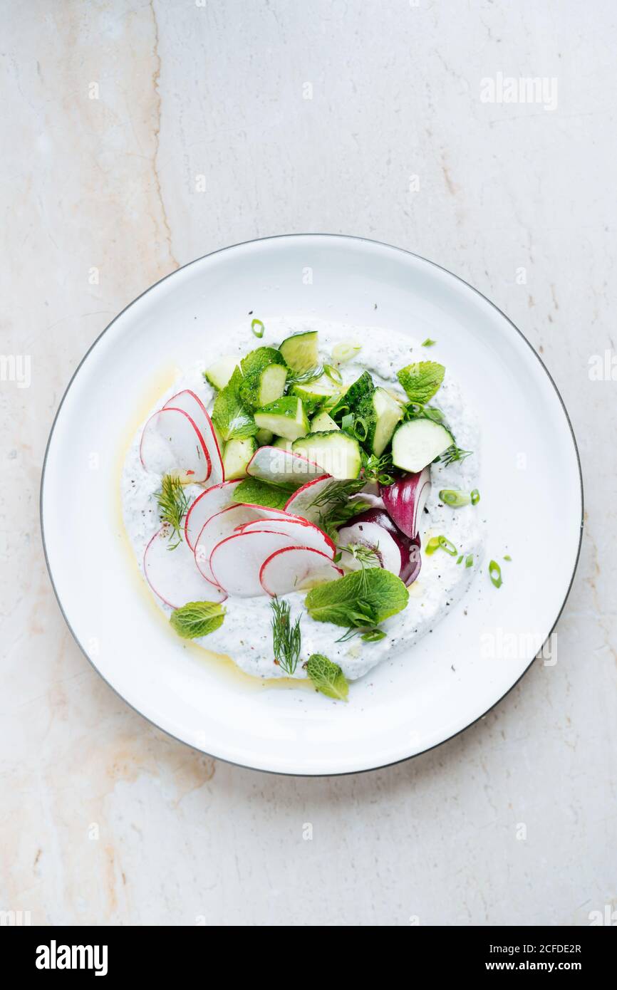 From above fresh salad of thin sliced radish ripe zucchini red onion and herbs with sour cream in white plate on table Stock Photo