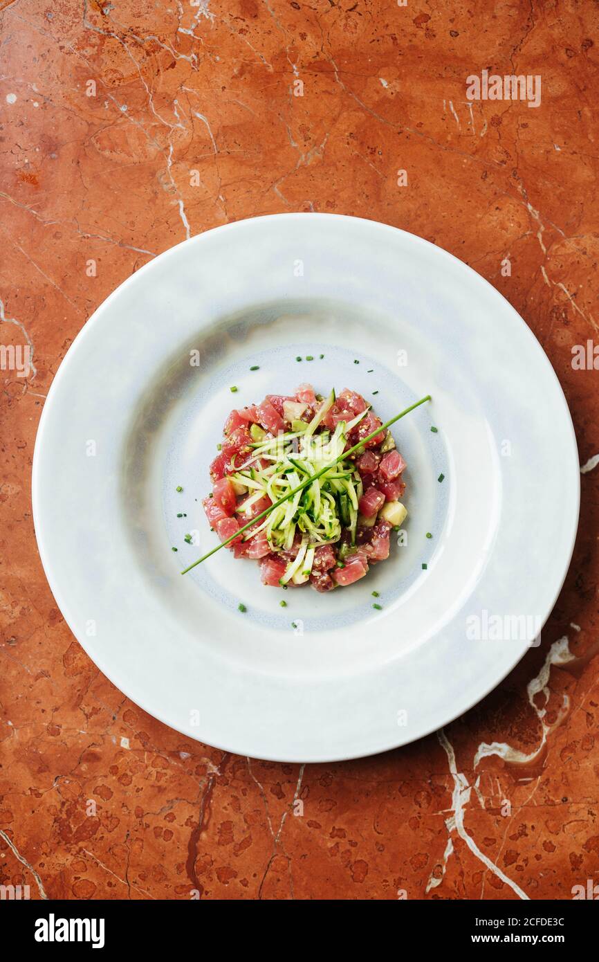 Top view of ceramic white plate with slices of red tuna and cheese decorated with pieces of cucumber and spices in restaurant Stock Photo