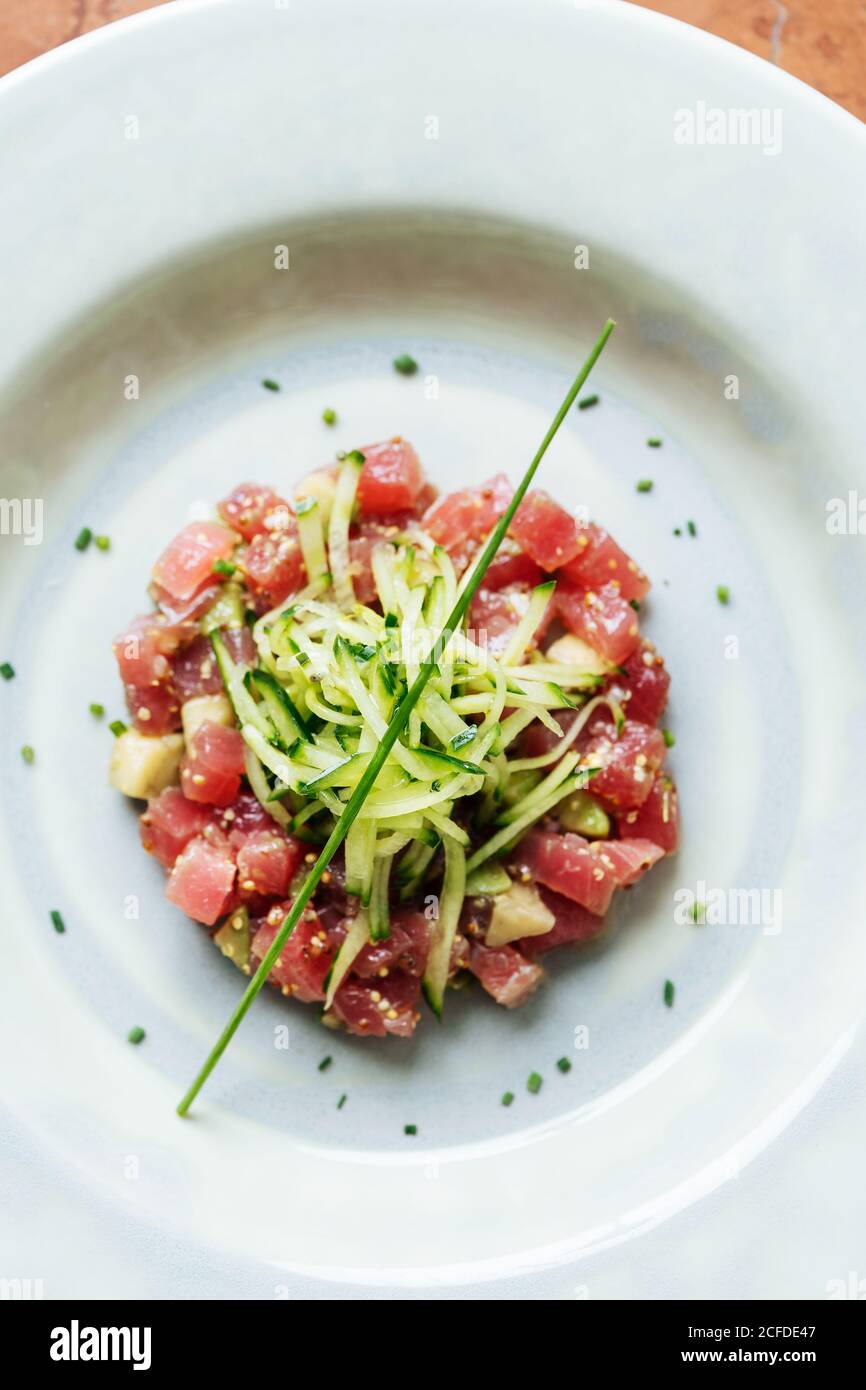 Top view of square pieces of red tuna served with slices of cucumber and herbs on white plate in restaurant Stock Photo