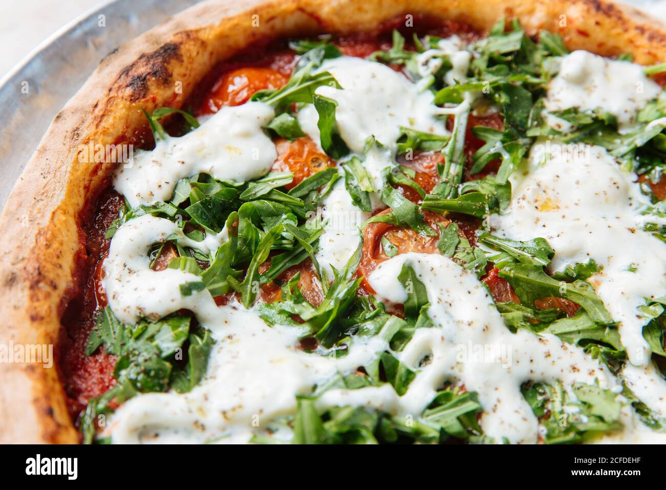 From above of thin baked pizza with thick edge filled with cherry tomatoes and chopped arugula with white ranch sauce topping Stock Photo