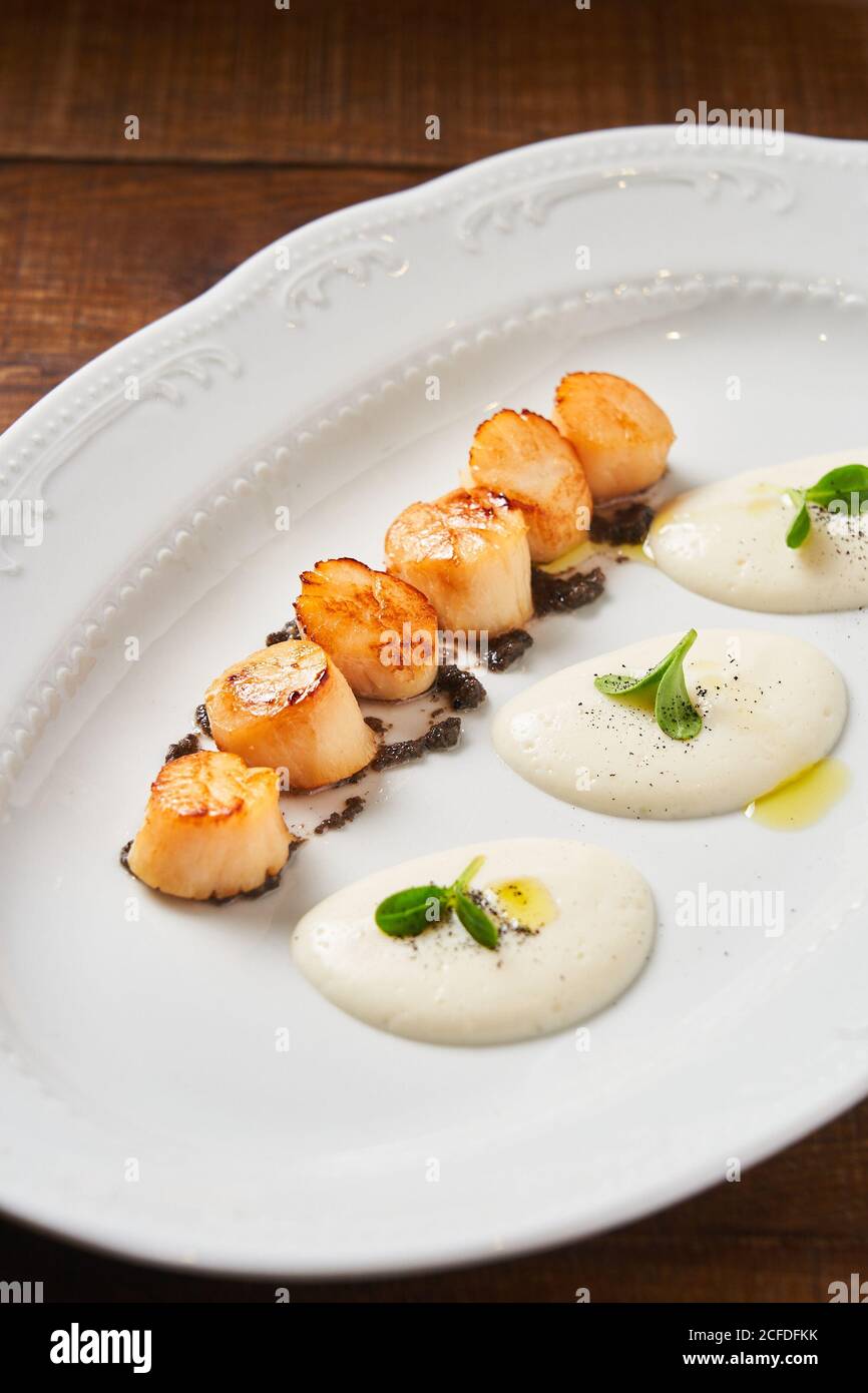 From above of fried scallops leading out on plate in row served with white creamy sauce and green herbs in restaurant Stock Photo