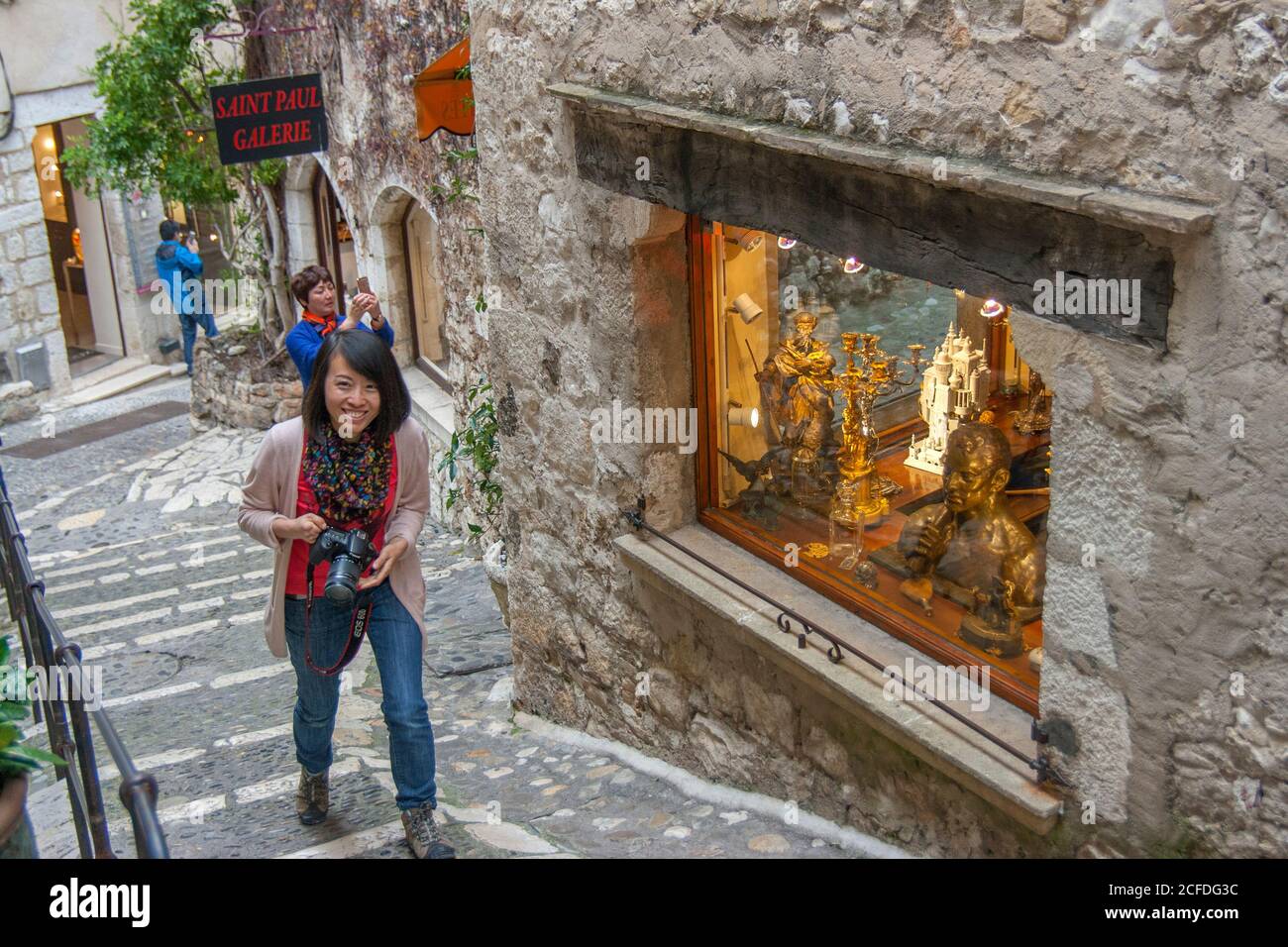 Saint-Paul-de-Vence France Photographers on every corner of the street. Smiling japanese tourist girl with camera. Artworks in the shop window. Stock Photo