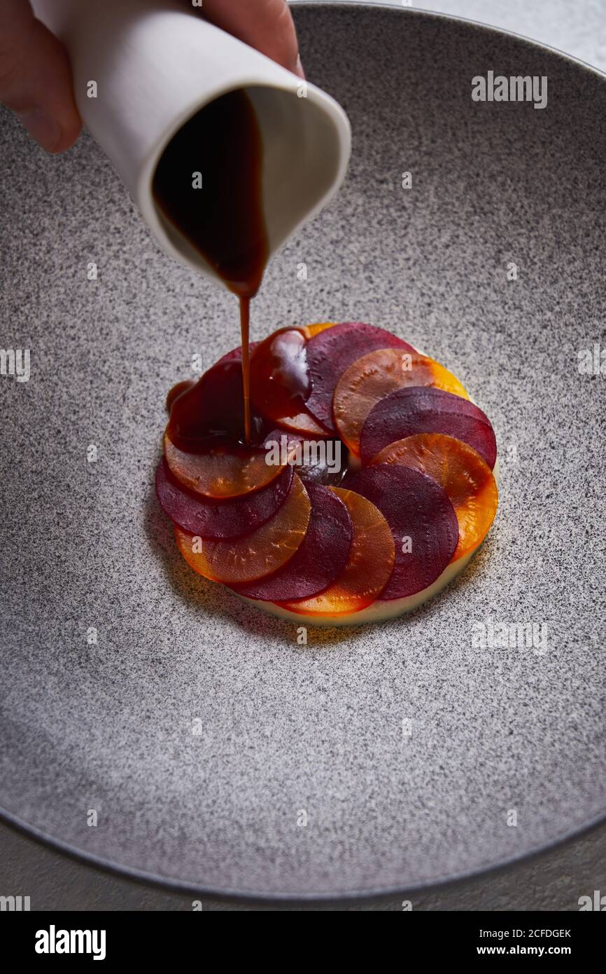 From above anonymous person adding sauce to delectable dish made of thin slices of boiled carrot and beetroot on plate Stock Photo