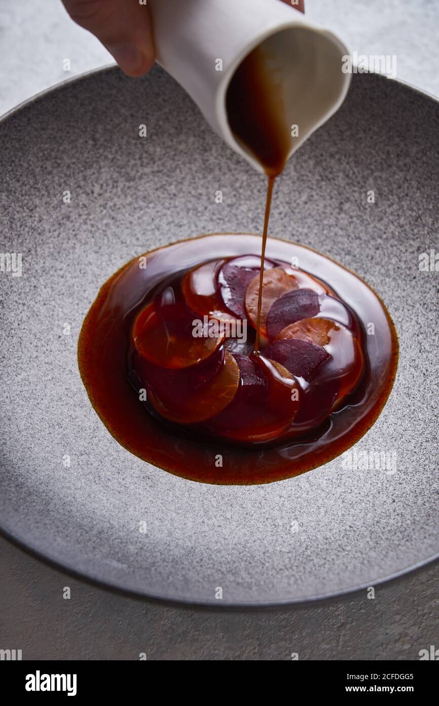 From above anonymous person adding sauce to delectable dish made of thin slices of boiled carrot and beetroot on plate Stock Photo