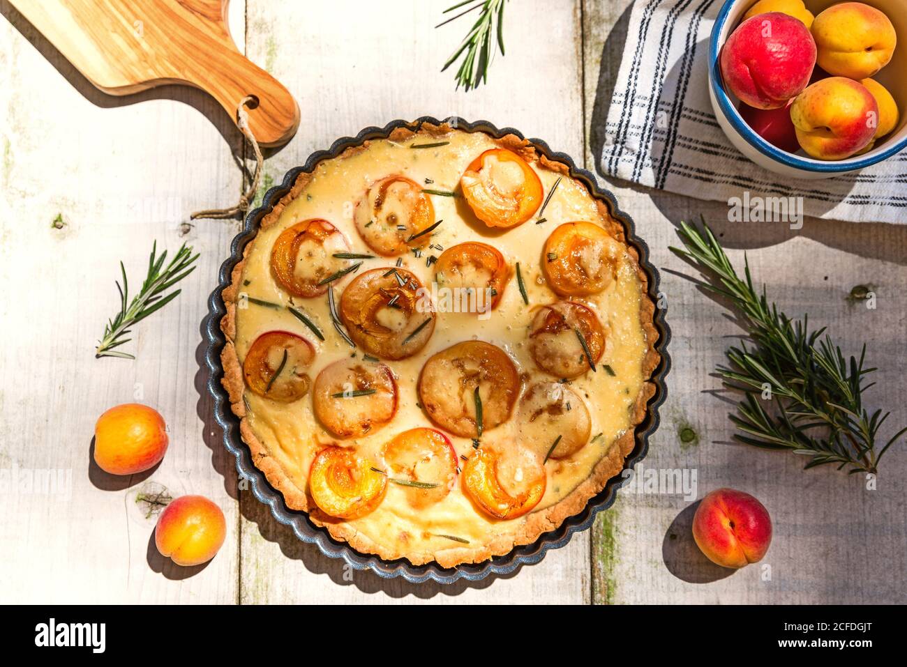 Apricot sour cream tart on a garden table with apricot rosemary decoration, bowl of apricots and cutting board cut at the edge of the picture. Stock Photo