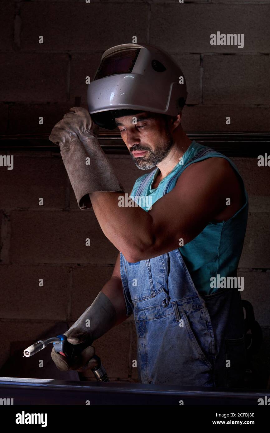 Professional man with welding mask on head wearing jeans overalls and protective gloves standing on workplace and preparing instrument for soldering while looking away Stock Photo
