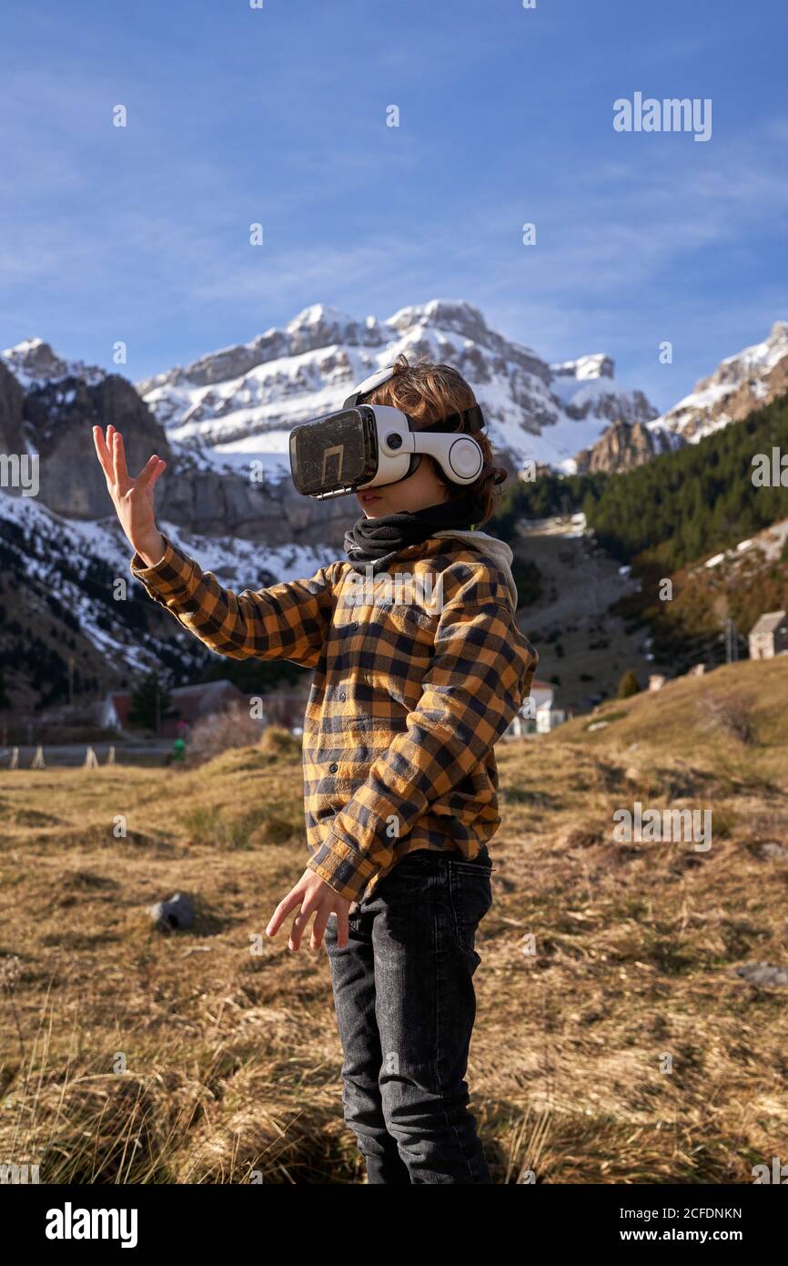 Active smart boy looking away in VR glasses standing on stone in mountain valley Stock Photo