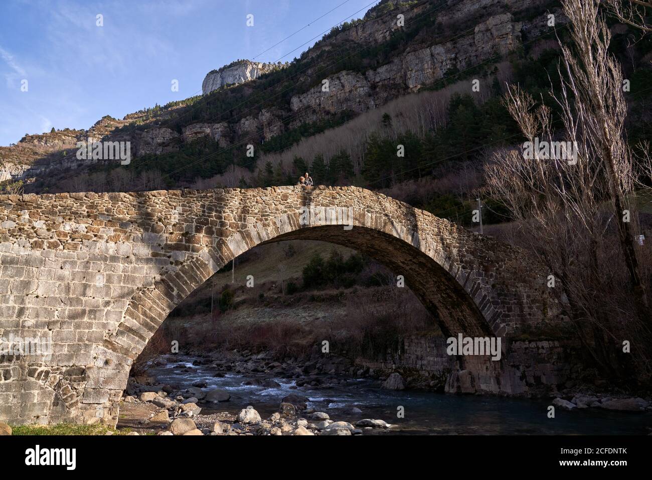 Woman and child appreciating landscape of ancient arched bridge in mountains crossing stream with dry leafless trees in bright day Stock Photo
