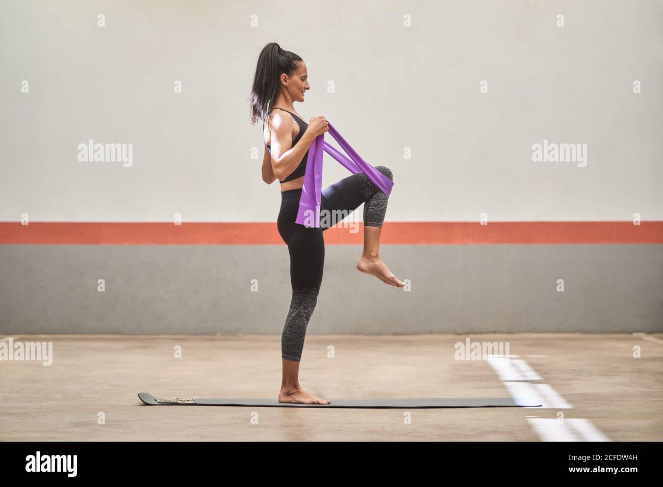 Cheerful athletic female in activewear standing barefoot on mat and stretching legs with resistance band during training Stock Photo