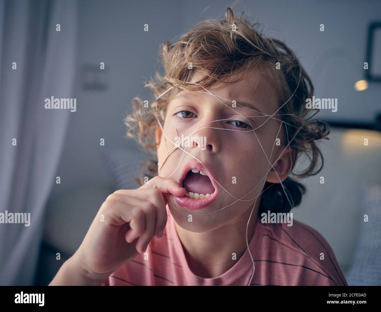 Naughty kid tangling in dental floss making faces while tiring floss to tooth to pull it out looking away Stock Photo
