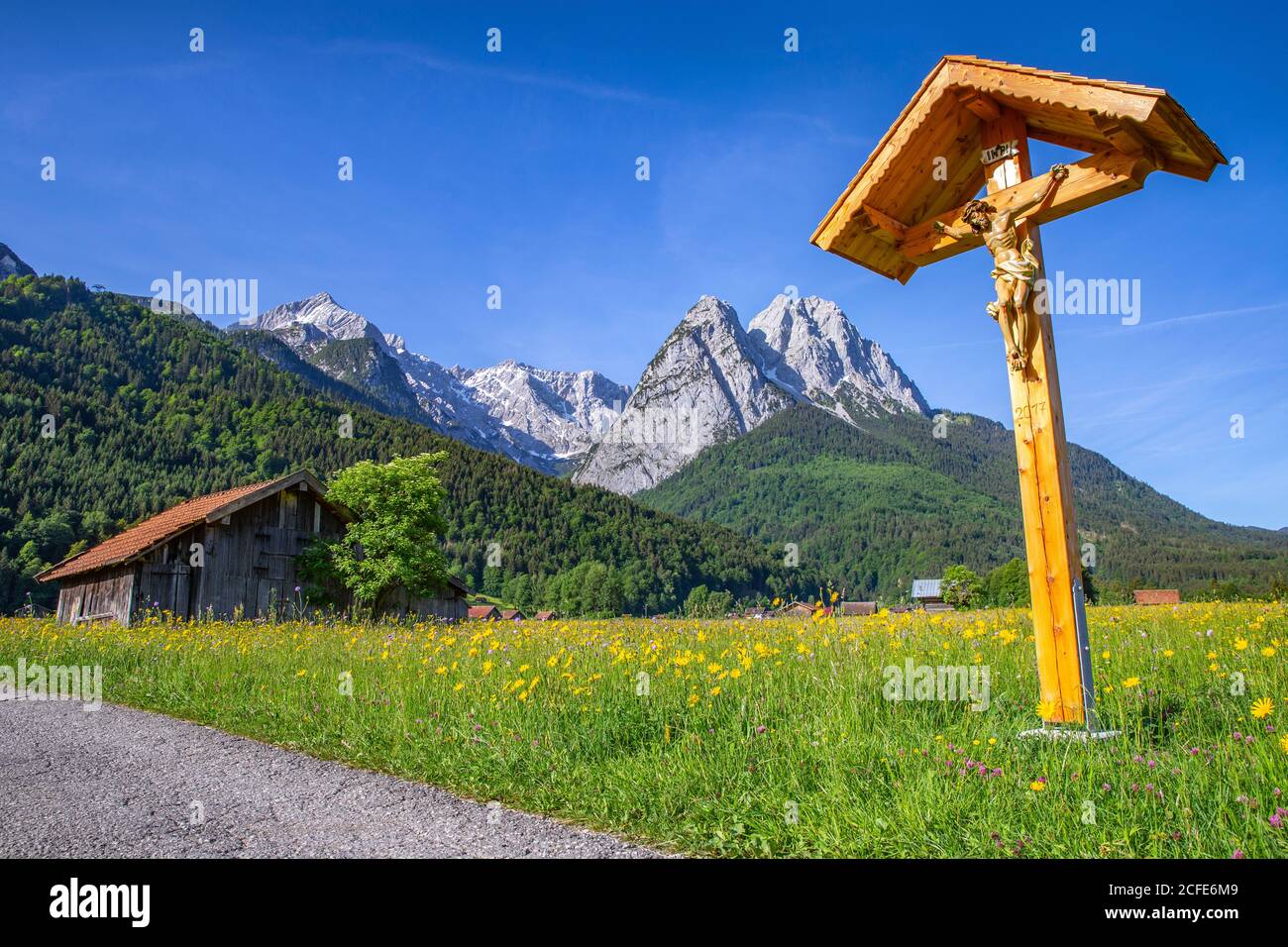 Flurkreuz (wayside cross, marterl, crucifix) at the field edge in spring near Garmisch-Partenkichen, view towards the Wetterstein Mountains with Stock Photo