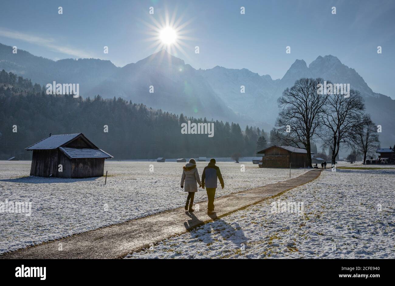 Man and woman on a winter walk on a dirt road in Garmisch-Partenkirchen, looking towards the Wetterstein Mountains with the Alpspitze and wax stones, Stock Photo