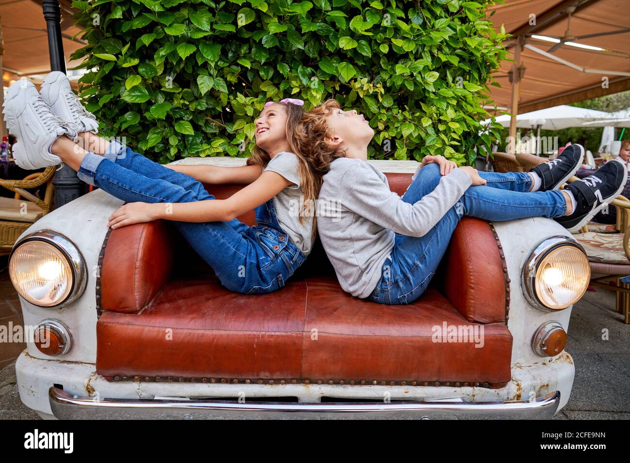 Side view of young lively children sitting back to back with legs up on car front converted into couch and looking upwards to ceiling while smiling and wondering Stock Photo