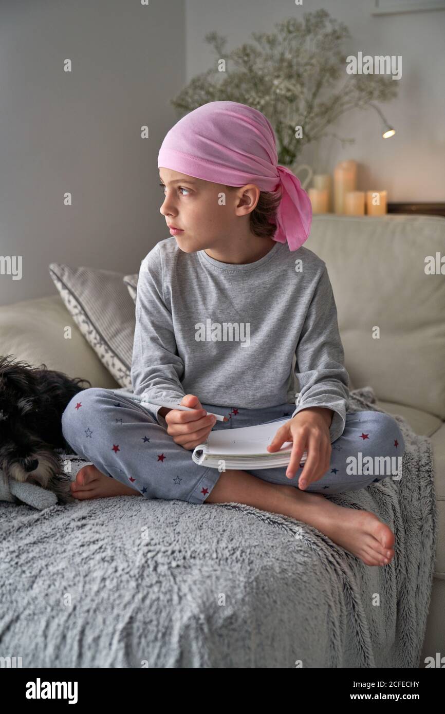 From below serious child with cancer diagnosis making notes while sitting on bed in room Stock Photo