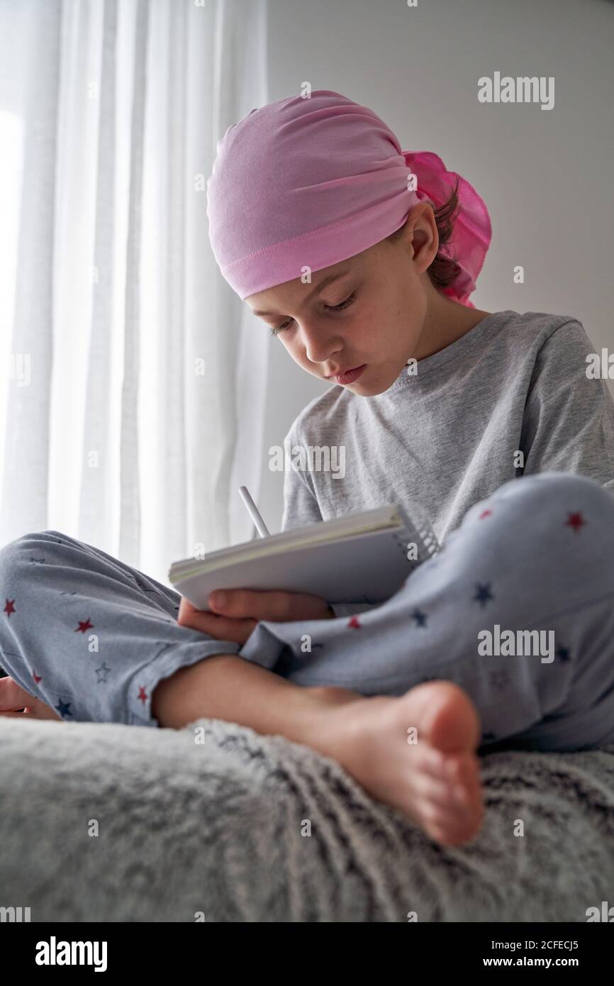 From below serious child with cancer diagnosis making notes while sitting on bed in room Stock Photo