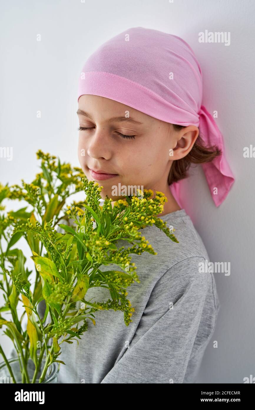 Focused little boy with cancer diagnosis wearing pink bandana with closed eyes while holding vase with flowers and standing at wall Stock Photo