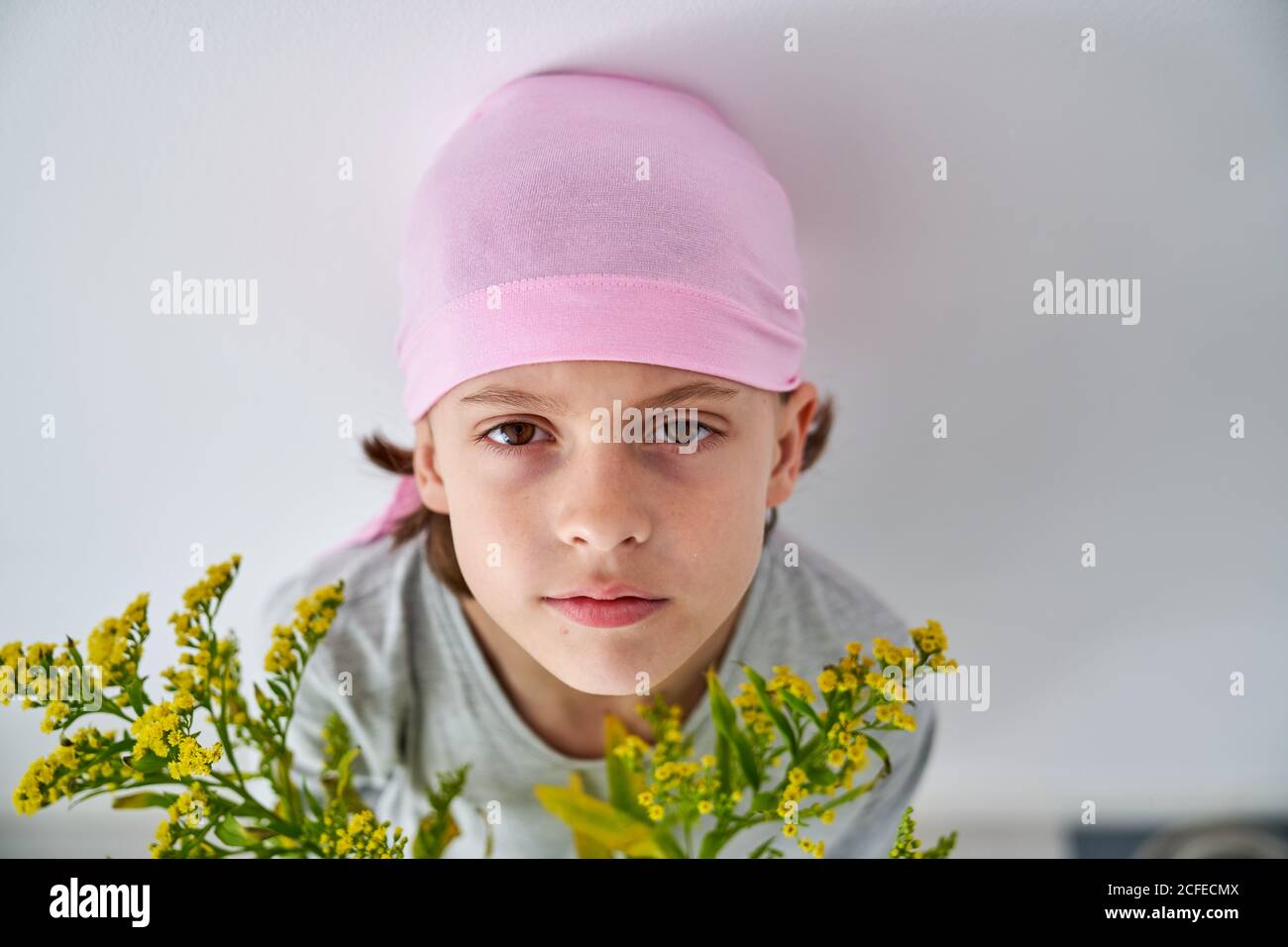 Focused little boy with cancer diagnosis wearing pink bandana and looking at camera while holding vase with flowers and standing at wall Stock Photo