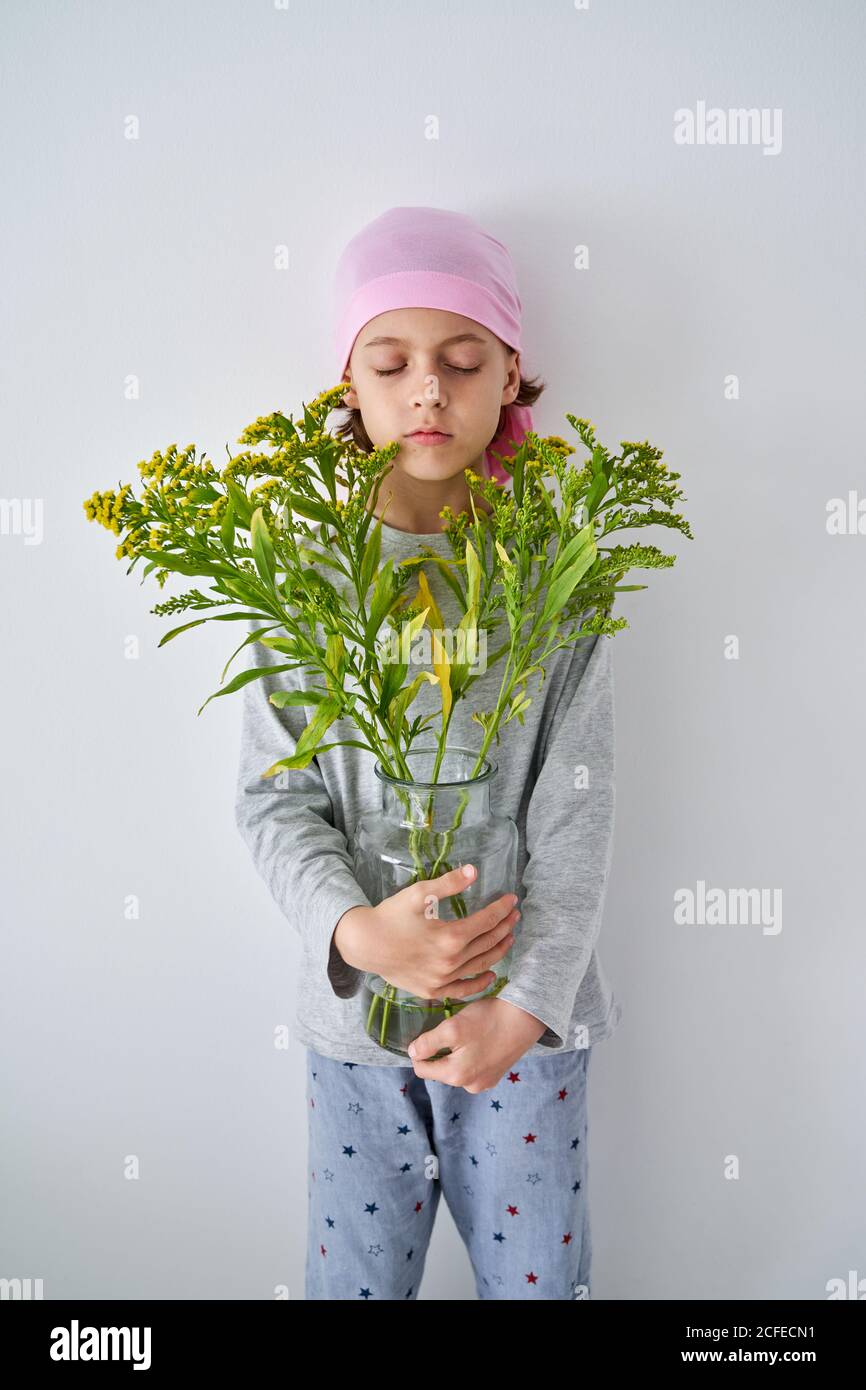 Focused little boy with cancer diagnosis wearing pink bandana with closed eyes while holding vase with flowers and standing at wall Stock Photo