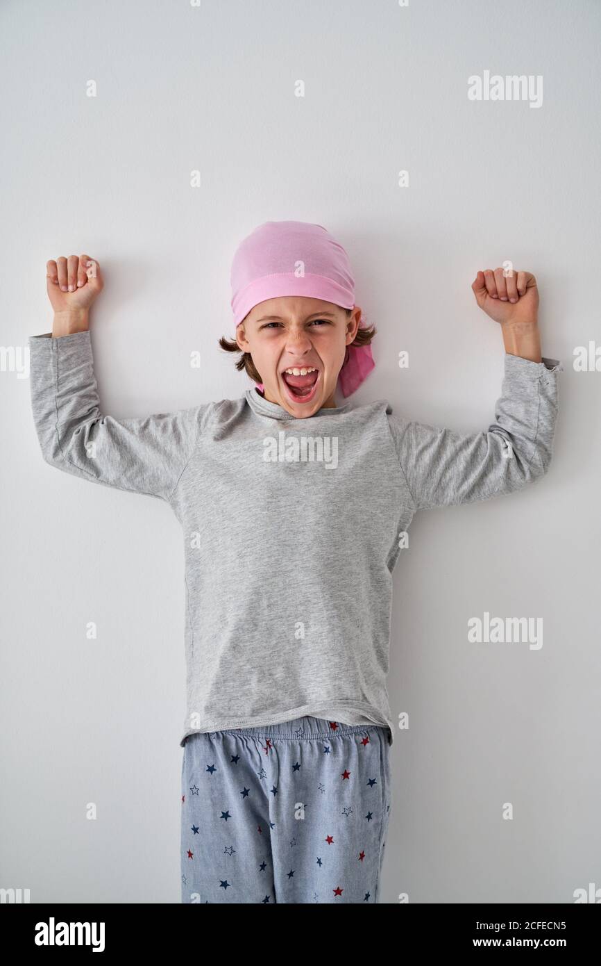 Brave small child with cancer diagnosis looking at camera and screaming while raising fists up on gray background Stock Photo