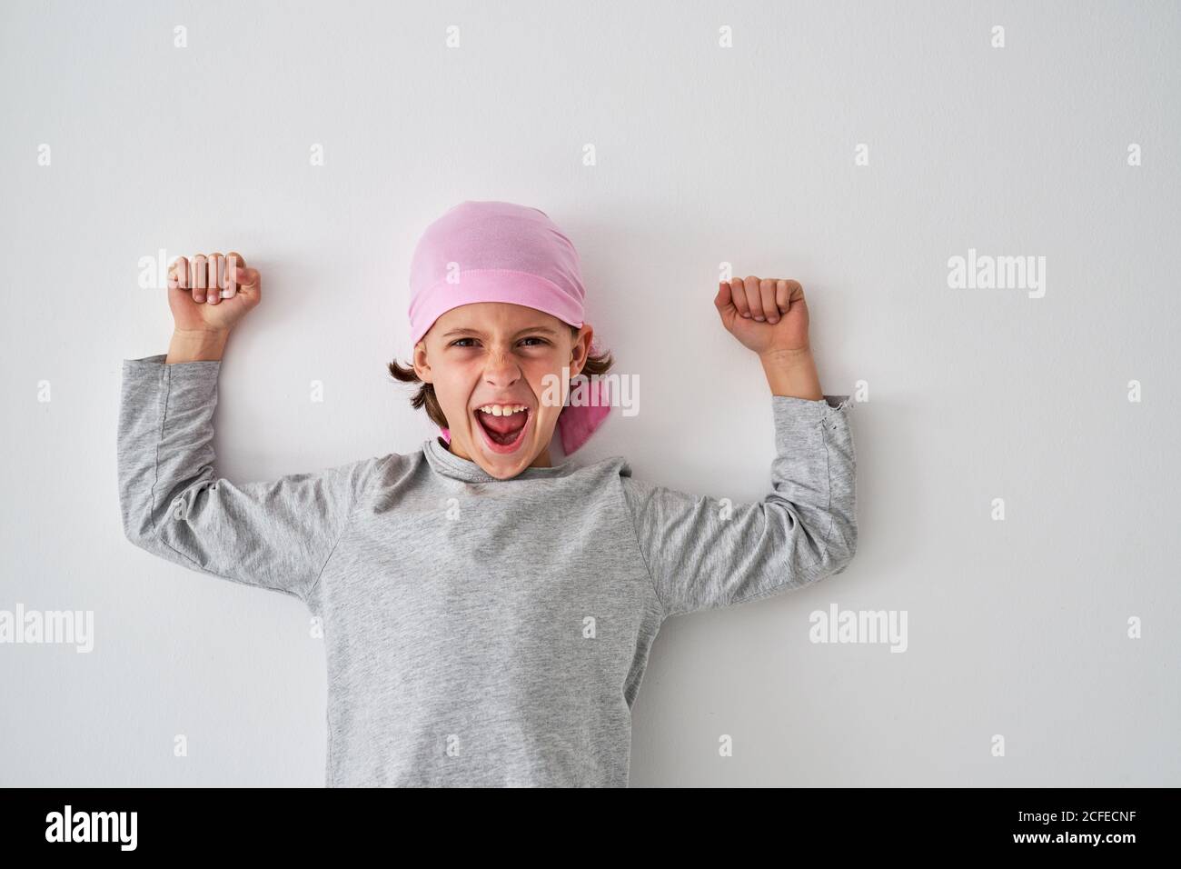 Brave small child with cancer diagnosis looking at camera and screaming while raising fists up on gray background Stock Photo
