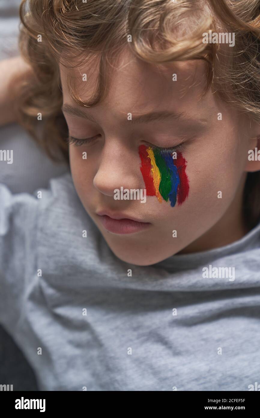 From above cute boy in pajama and with rainbow under eye lying on pillow and blanket and sleeping while staying home during quarantine Stock Photo