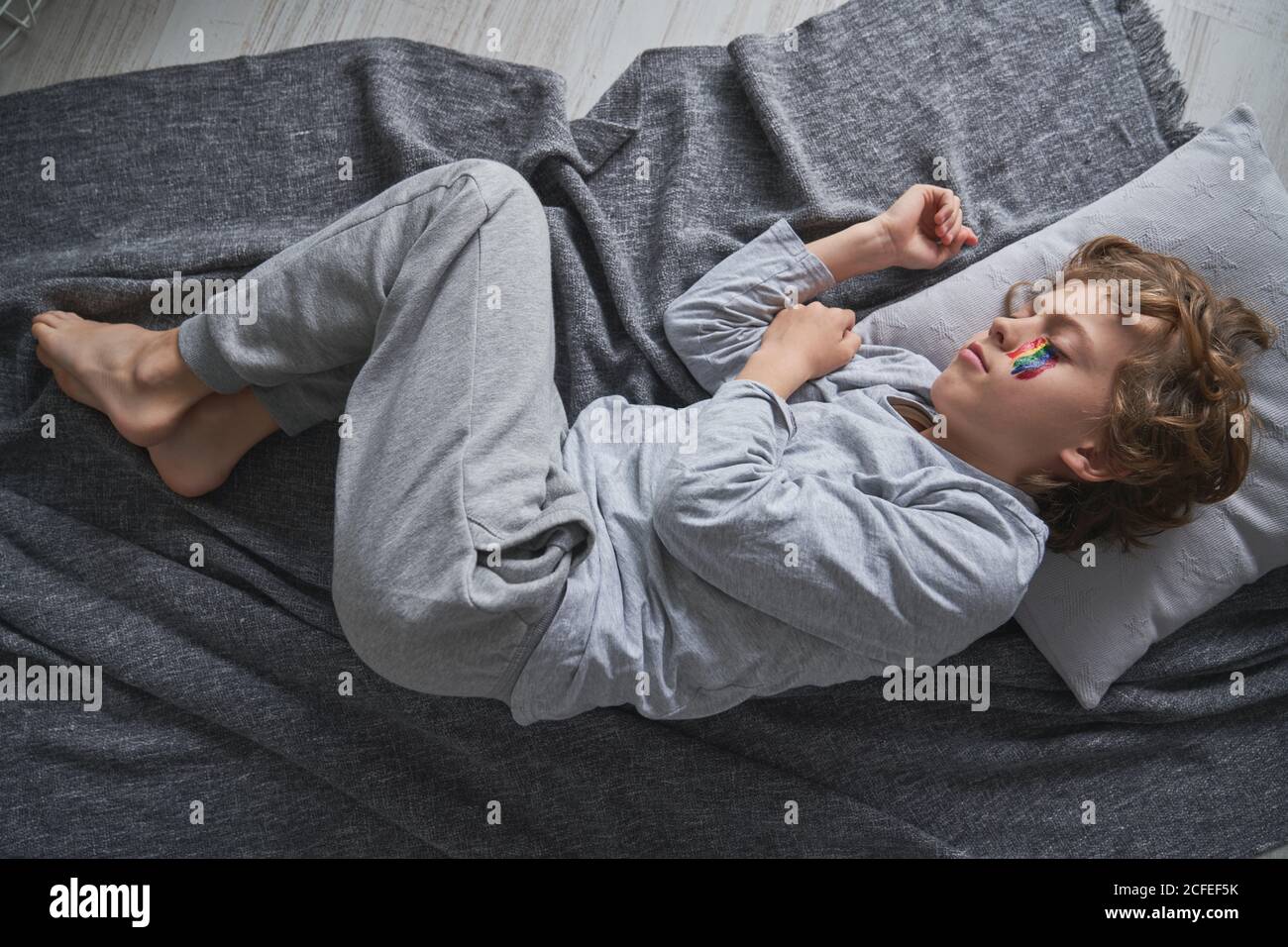 From above cute boy in pajama and with rainbow under eye lying on pillow and blanket and sleeping while staying home during quarantine Stock Photo