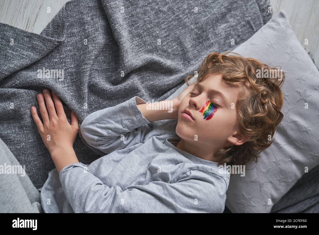 From above cute boy in pajama and with rainbow under eye lying on pillow and blanket and sleeping while staying home during quarantine Stock Photo