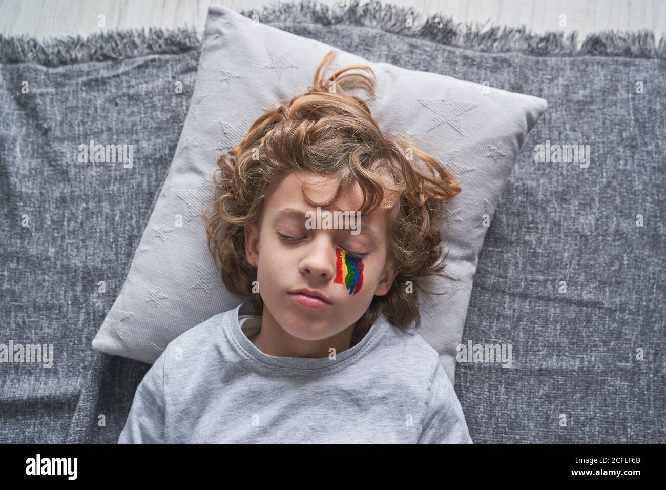 From above cute boy in pajama and with rainbow under eye lying on pillow and blanket and sleeping while staying home during quarantine Stock Photo
