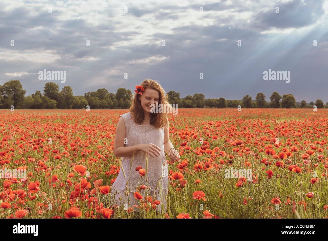 Young girl with reddish hair stands in the huge red poppy field and smiles carefree Stock Photo