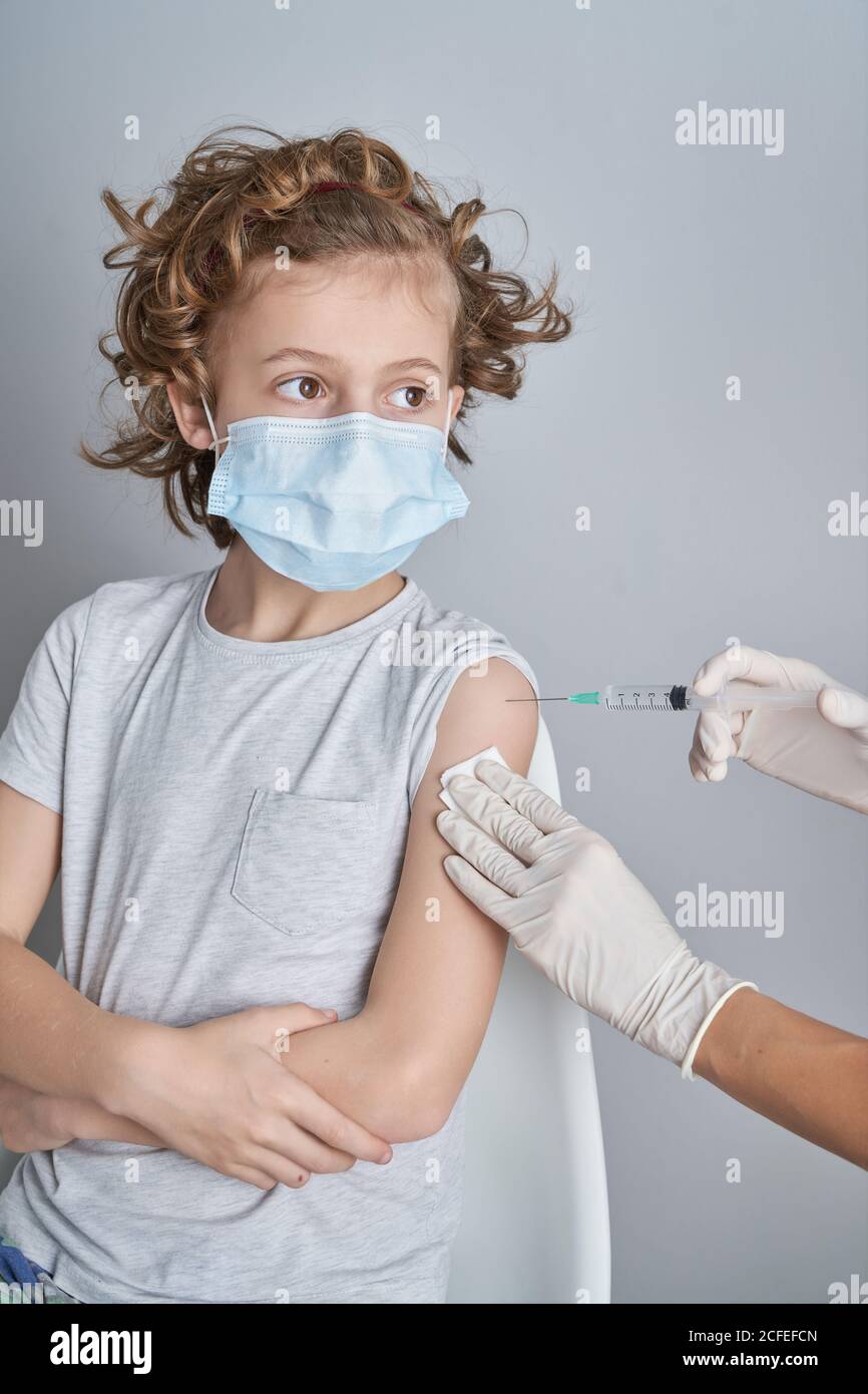 Crop nurse in white latex gloves holding shoulder of boy with curly hair while giving vaccine injection with syringe in modern clinic Stock Photo