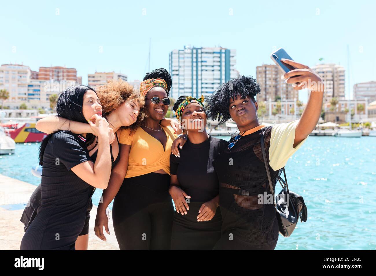 Group of charismatic multiracial girlfriends standing at waterfront and taking selfie on smartphone during stroll in summer Stock Photo