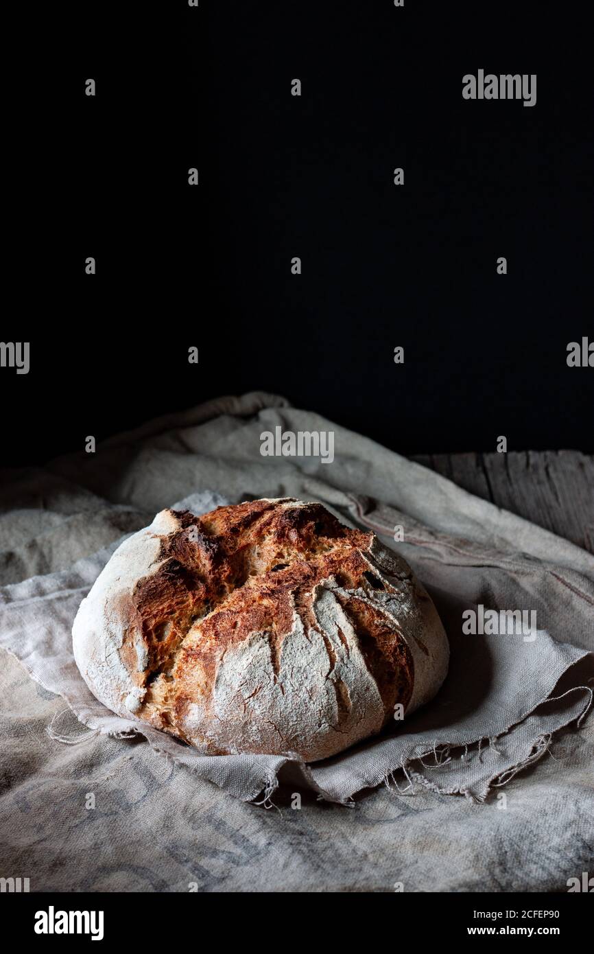 Loaf of fresh country sourdough bread placed on piece of wood on shabby table against black background Stock Photo