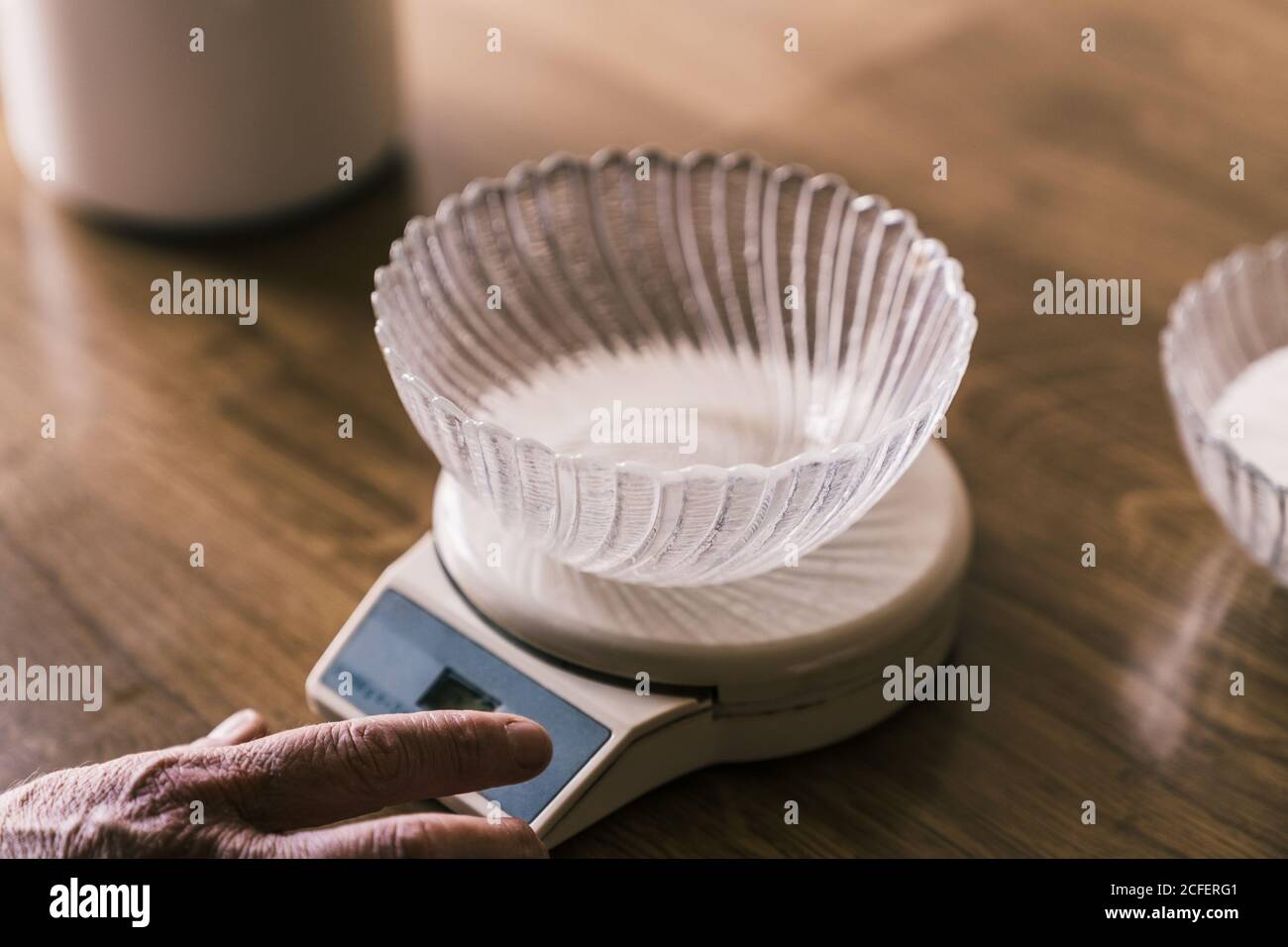 Cropped unrecognizable female hand weighting with electronic kitchen scale with glass bowl on top on wooden table Stock Photo