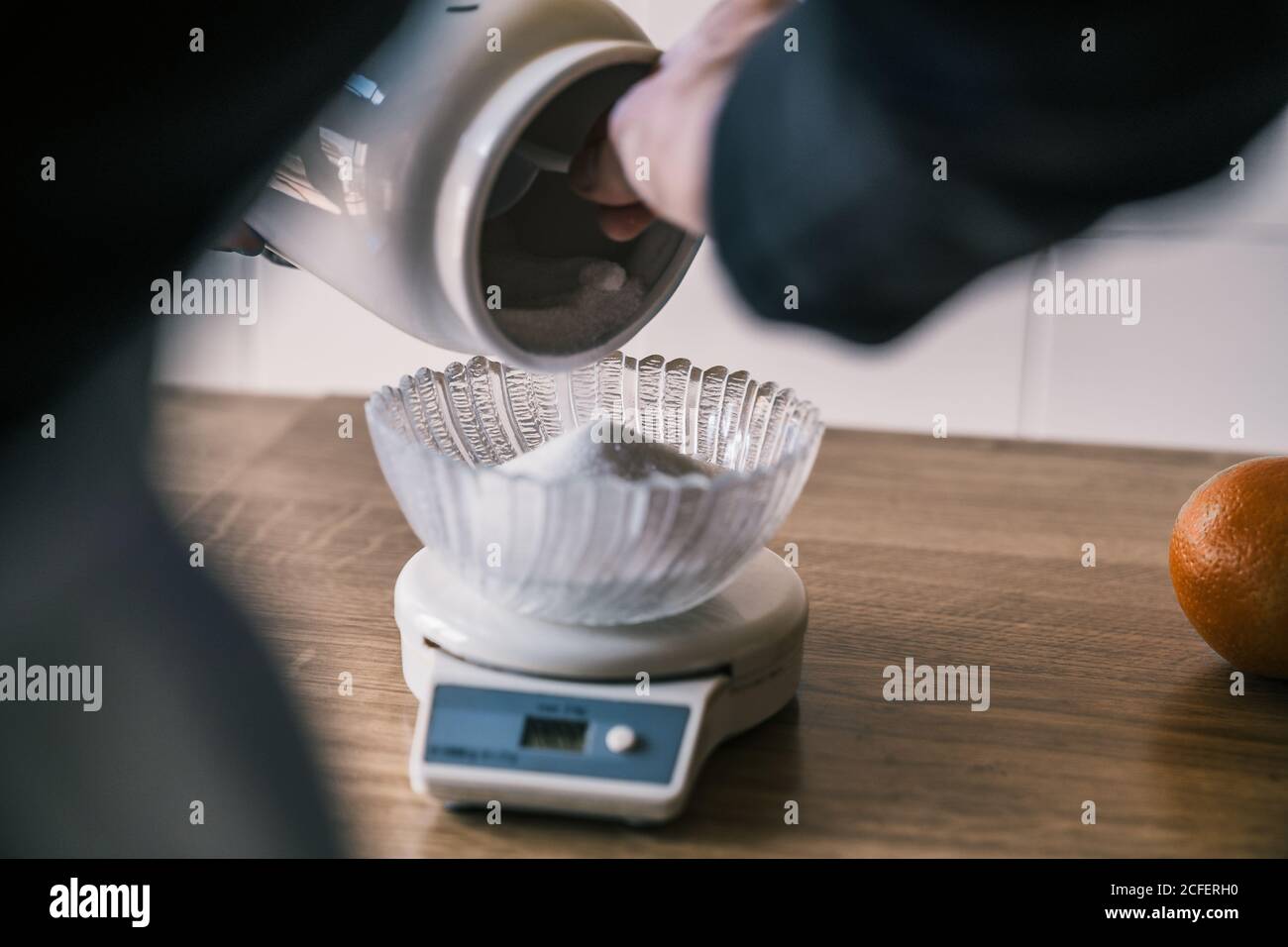 Cropped unrecognizable female hand holding sugar jar spilling and weighting sugar with electronic kitchen scale with glass bowl on top on wooden table Stock Photo