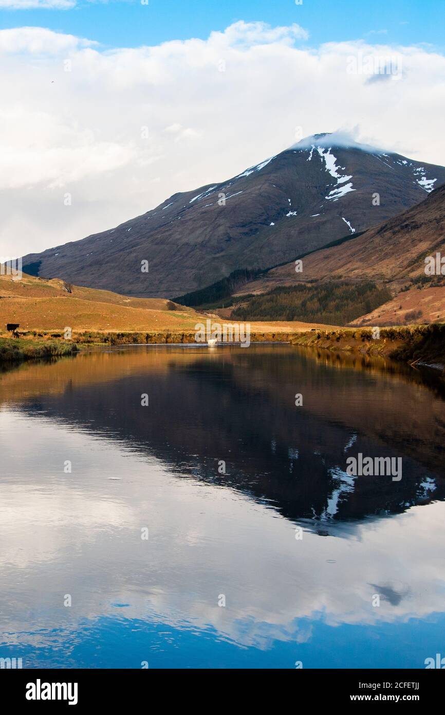 Amazing Scottish landscape of calm lake with mirrored surface reflecting mountain with snow covered peak and blue cloudy sky in Glen Coe area Stock Photo