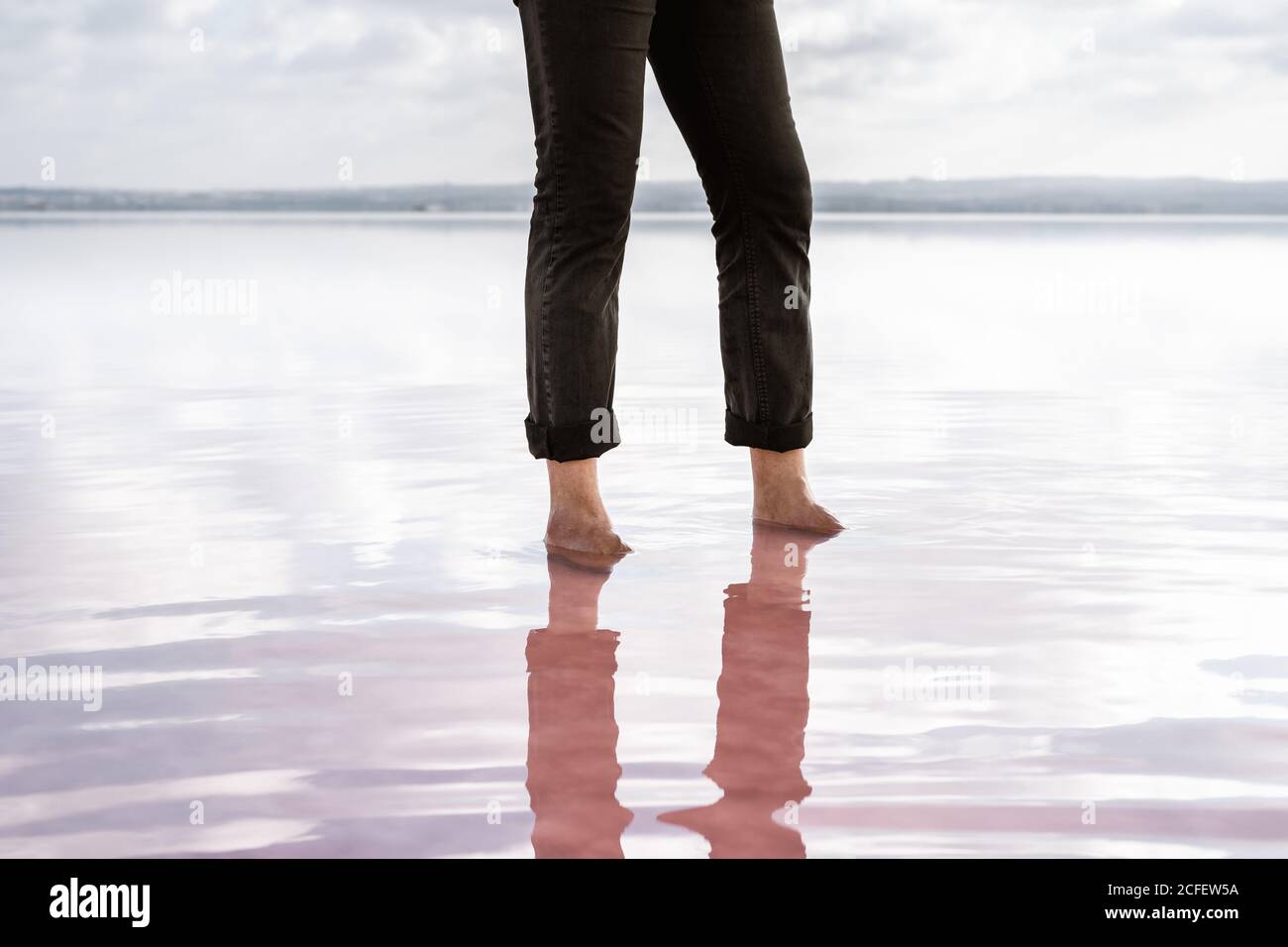 Crop barefoot man in black pants standing in still sea by shore on cloudy day Stock Photo