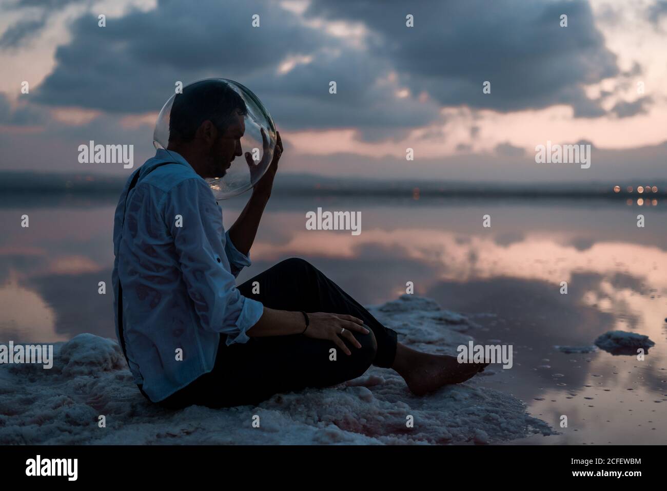 Pensive man with closed eyes in wet shirt taking out empty aquarium on head while sitting still by the seaside in twilight Stock Photo
