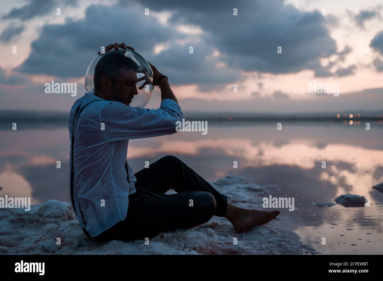 Pensive man with closed eyes in wet shirt taking out empty aquarium on head while sitting still by the seaside in twilight Stock Photo