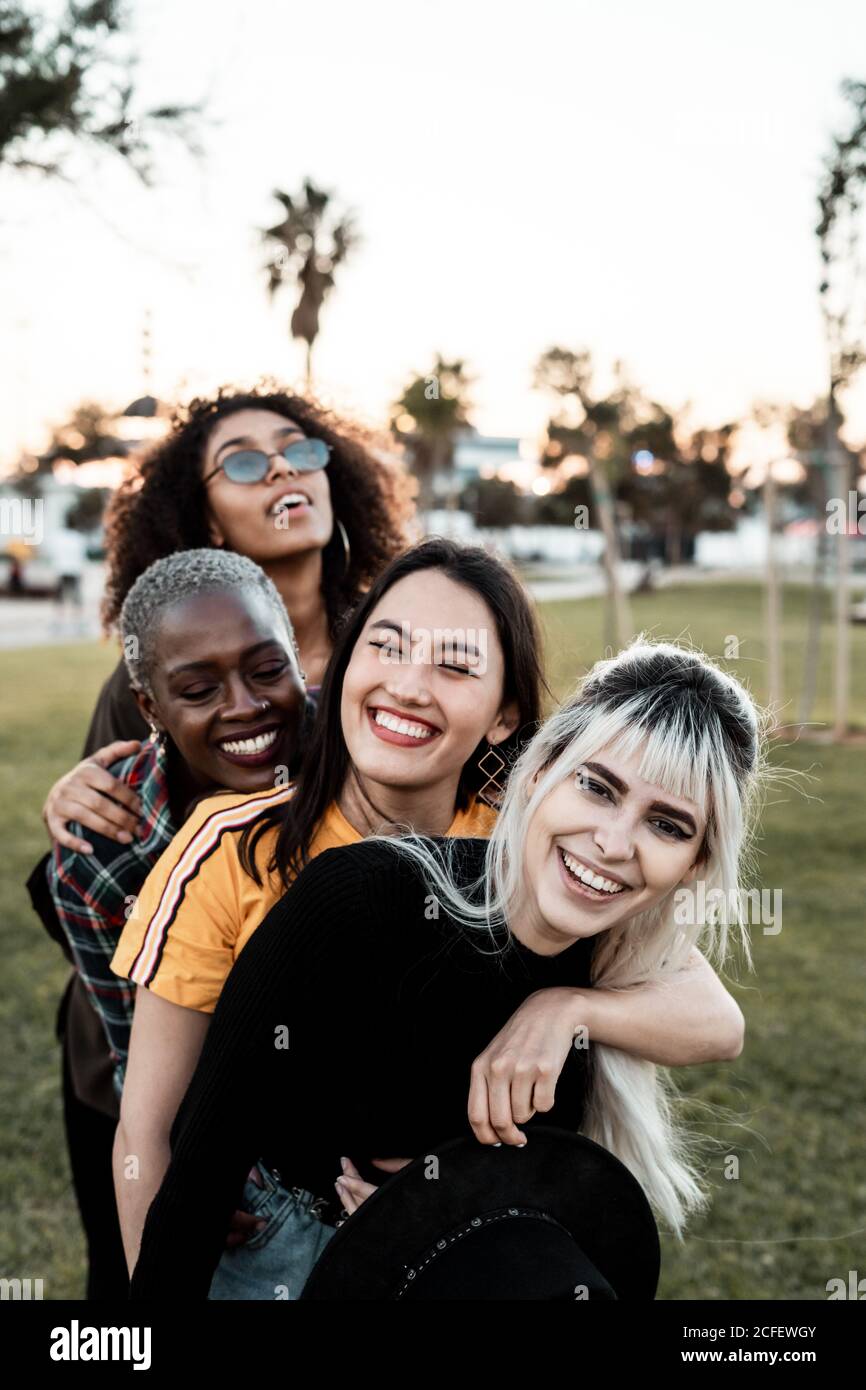 Multiethnic group of young laughing female friends in casual clothes having fun and cuddling with each one on lawn Stock Photo