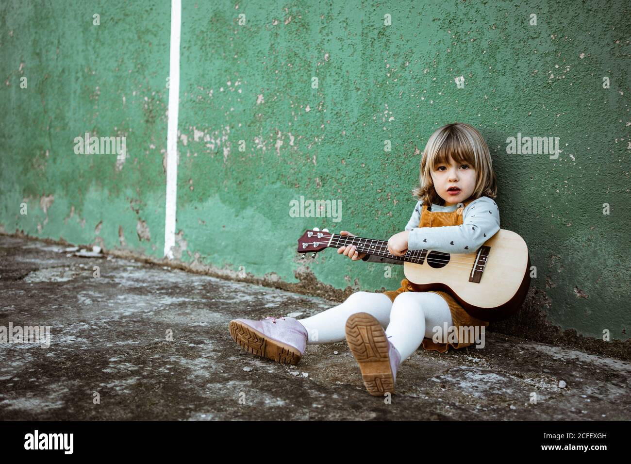 Little girl with ukulele sitting on rough ground near kick scooter against weathered green wall on street Stock Photo
