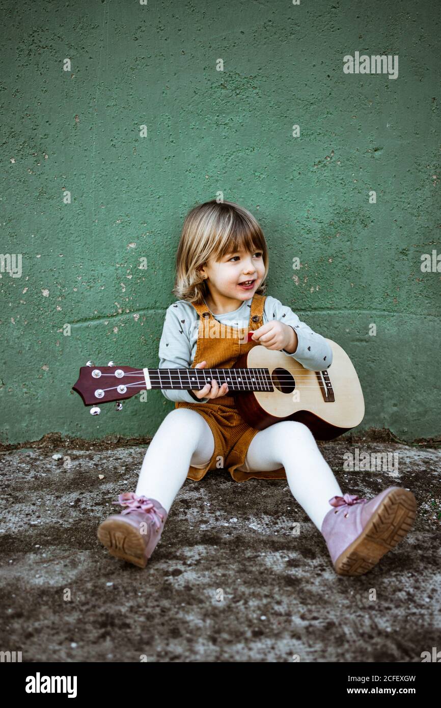Little girl with ukulele sitting on rough ground near kick scooter against weathered green wall on street Stock Photo