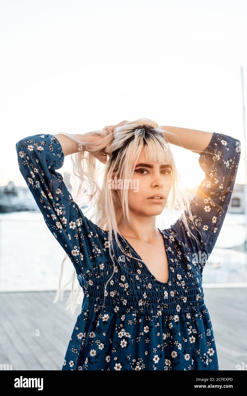 Gorgeous pensive blond female with black eyebrows in blue oversize dress looking at camera with curiosity while standing and touching hair on street during sunset Stock Photo