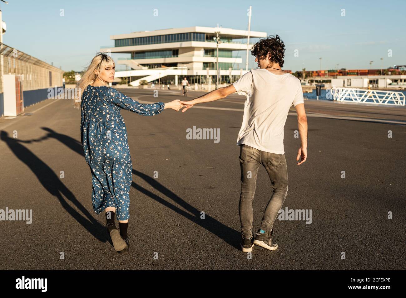 Back view of pensive blond walking Woman in blue oversize dress looking back over shoulder and holding hand of handsome man in casual clothes going away Stock Photo
