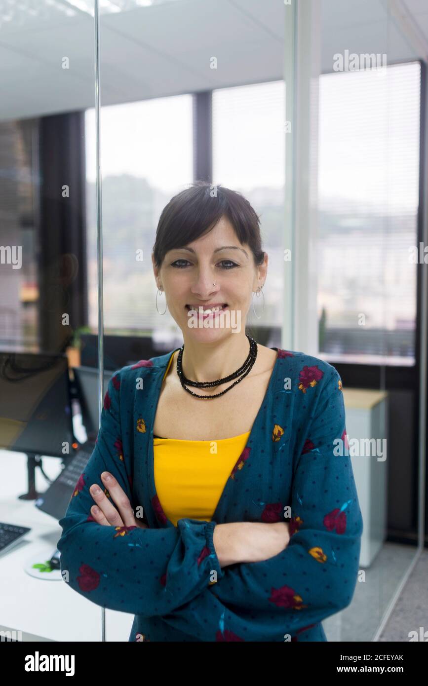 Attractive adult female keeping eyes crossed and smiling while standing near glass wall in