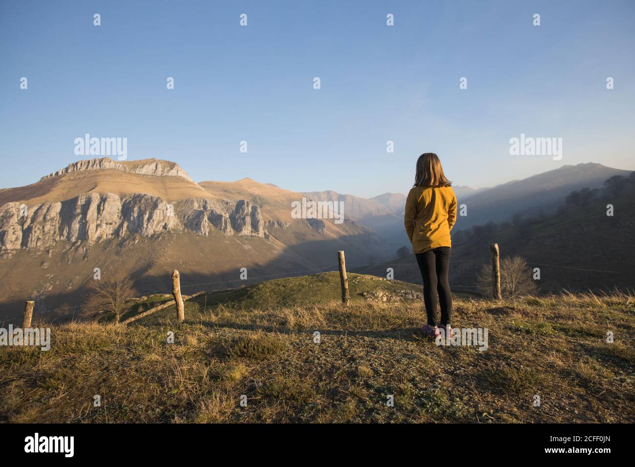 Back view of faceless girl in casual clothes standing alone on green hill and enjoying picturesque view of mountains and sky in Cantabria Stock Photo
